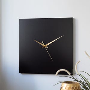 May include: A square, black wall clock with gold hands. The clock has a minimalist design and is mounted on a white wall. The clock's hands indicate a time of approximately 1:10.