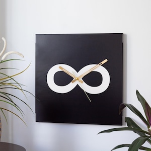 May include: Square black wall clock featuring a white infinity symbol. The clock hands are gold-colored. The clock is mounted on a white wall, with plants and a decorative vase in the foreground. A modern home decor item.