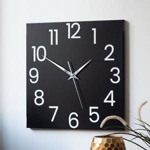 Może przedstawiać: Czarny kwadratowy zegar z białymi cyframi i srebrnymi wskazówkami. Zegar wisi na białej ścianie.
