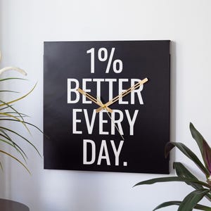 May include: Square black wall clock with gold-colored hands. The clock face features the text "1% BETTER EVERY DAY" in white, a motivational message. The clock is hanging on a white wall, with green plants in the foreground.
