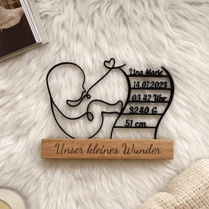 May include: A black metal art piece depicting a baby in a womb-like shape, mounted on a wooden base. The piece includes the name "Tina Marie", date, time, weight (3240 g), and length (51 cm). The text "Unser kleines Wunder" is engraved on the wooden base.