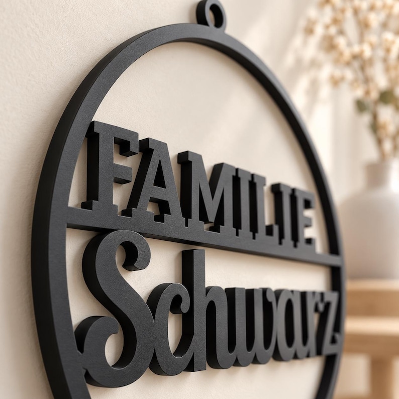 May include: A circular black wall sign featuring the word FAMILIE in a bold, block-style font on the top half and the word Schwarz in a cursive script font on the bottom half, with a small circular loop at the top for hanging purposes.