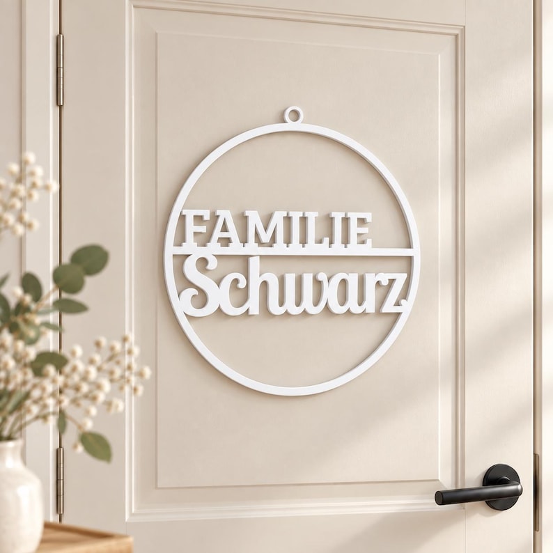 May include: A white circular wall sign featuring a small hanging loop at the top. The sign contains the text "FAMILIE" in a bold, sans-serif font on the upper half and the word "Schwarz" in a decorative, cursive script on the lower half.