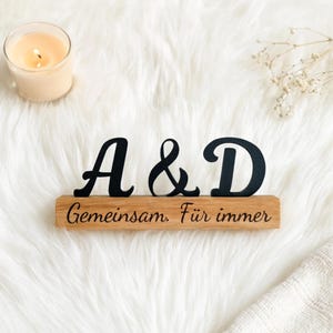 May include: A wooden sign featuring the letters "A & D" in black script, with the engraved words "Gemeinsam. Für immer". A lit candle and decorative flowers are visible in the background.