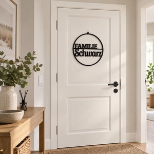 May include: A circular black metal wall sign featuring the text FAMILIE Schwarz in a bold, sans-serif font. The sign is mounted on a white interior door with a black handle. The design includes a thin circular frame with a small hanging loop at the top.