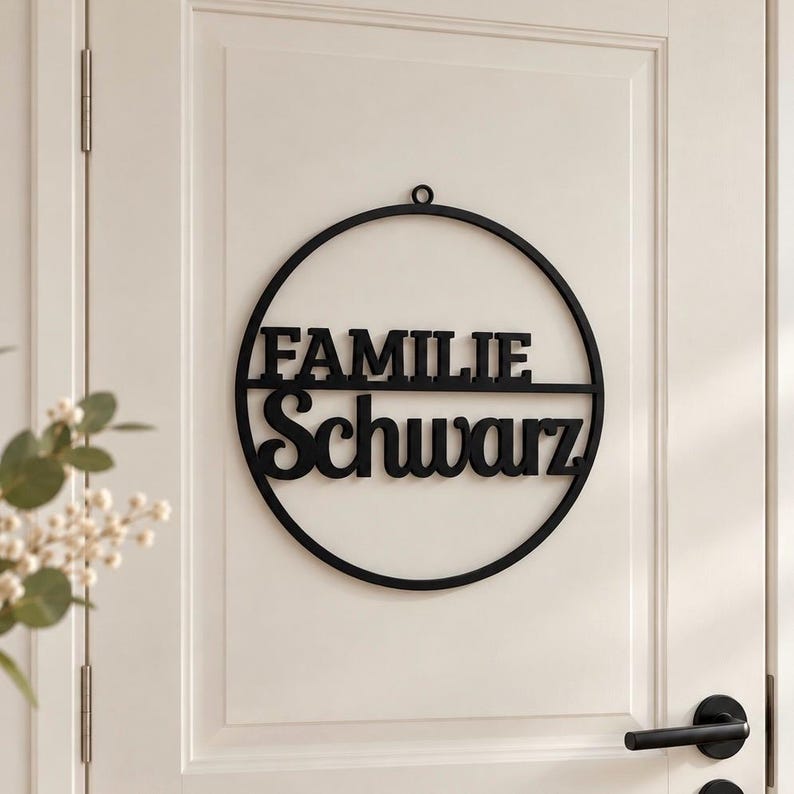 May include: A circular black wall sign featuring the word FAMILIE in a sans-serif font on the top half and the word Schwarz in a script font on the bottom half, with a small loop at the top for hanging, mounted on a white paneled door.