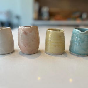 May include: Four small ceramic creamer jugs in different colours: white, pink, yellow, and blue. The jugs are all slightly different shapes and have a rustic, handmade look.