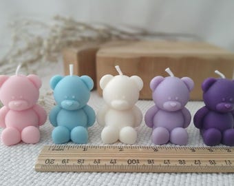 May include: Five bear-shaped candles in pastel colors: pink, blue, white, purple, and dark purple. Each candle has a wick on top and is approximately 2 inches tall. A wooden ruler is at the bottom.