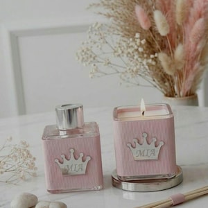 May include: A pink glass fragrance diffuser and a candle with a silver crown design and the name "MIA". The candle is lit, and the diffuser has a silver cap. Decorative dried flowers are in the background.