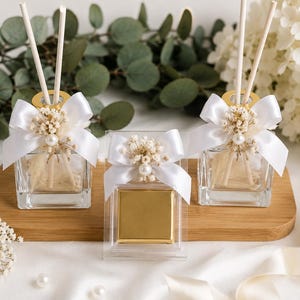 May include: Two square glass reed diffusers and one square gold-wrapped chocolate bar in a clear box. Each item is decorated with a white satin ribbon bow, a cluster of dried baby's breath flowers, and a single white pearl bead accent.
