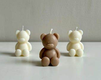 May include: Three teddy bear-shaped candles. Two are ivory, and one is brown. Each candle has a wick at the top. The bears are sitting on a white surface.