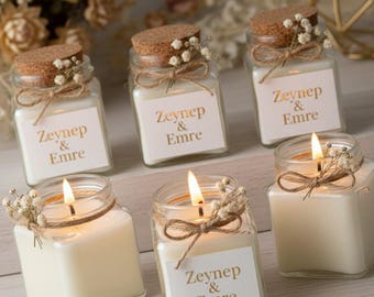May include: Five square glass jars filled with white wax candles. Each jar has a cork lid, a twine bow, and a small sprig of dried white flowers. A white label on each jar features the gold text Zeynep & Emre. Three jars are closed, and two are lit.