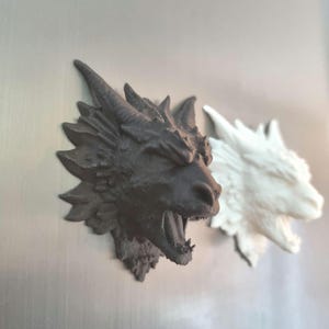 May include: Two dragon head sculptures, one dark brown and one white, are mounted on a light grey surface. The dragon heads have open mouths and textured scales, showcasing a fantasy theme.