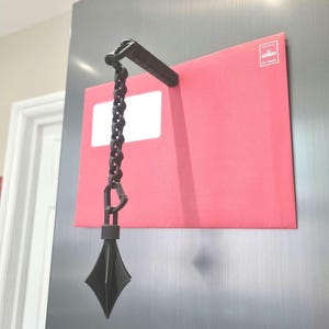 May include: A black envelope holder with a chain and a diamond-shaped weight. The holder is attached to a red envelope with a white window and a small label. The holder is hanging on a silver surface.