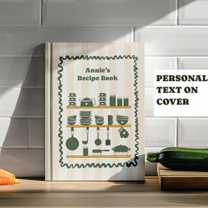 May include: A personalized recipe book with the text "Annie's Recipe Book" on the cover. The cover features a green and beige design with illustrations of kitchen utensils and dishes. The book is on a wooden surface with carrots and zucchini.
