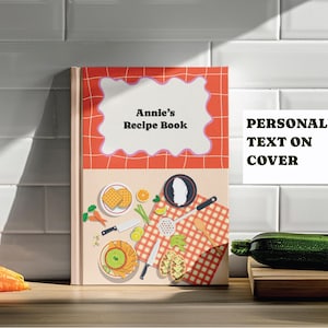 May include: A personalized recipe book titled "Annie's Recipe Book" with a red and white design. The cover features illustrations of food items like waffles, carrots, and a cutting board. The text "PERSONALIZED TEXT ON COVER" is also visible.