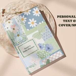 May include: A floral-patterned daily planner with a patchwork design featuring daisies, butterflies, and other flowers in pastel colors. The planner has the text "Annie's Daily Planner" on the cover and the words "PERSONALIZED TEXT ON COVER/SPINE" are also visible.