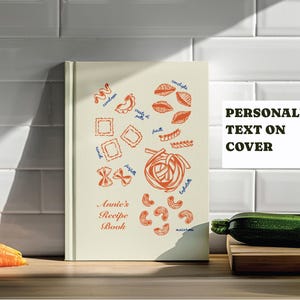 May include: A cream-colored recipe book with pasta illustrations in orange and blue. The book cover features various pasta shapes and the text "Annie's Recipe Book." The words "Personalized Text on Cover" are also visible.