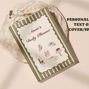May include: A personalised daily planner with a cream cover. The cover features the text "Annie's Daily Planner" in a decorative font, a scalloped border, and illustrations. The spine has green and white stripes.