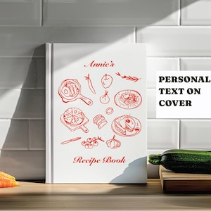 May include: A white recipe book with red illustrations of food items, including a pizza, fried egg, and apple. The cover reads "Annie's Recipe Book" with the text "PERSONALIZED TEXT ON COVER" in a white rectangle.