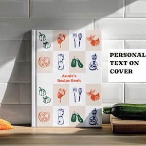Puede incluir: Un libro de recetas personalizado con un patrón de cuadros de ilustraciones de alimentos. La portada dice "Annie's Recipe Book" y "Texto personalizado en la portada". Zanahorias y calabacín son visibles.