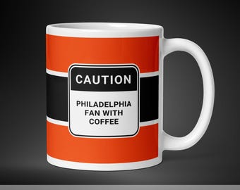 Pode incluir: Caneca de café de cerâmica branca com um design vermelho, preto e branco. A caneca tem um sinal de advertência que diz "ATENÇÃO FÃ DE FILADÉLFIA COM CAFÉ".