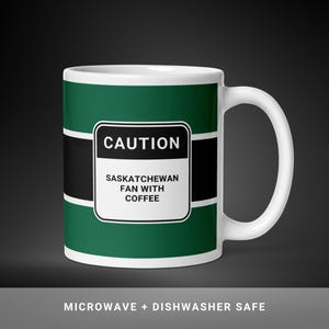 May include: White ceramic mug with a green and black design. The mug features the text "CAUTION SASKATCHEWAN FAN WITH COFFEE" in a white box. The bottom of the mug has the text "MICROWAVE + DISHWASHER SAFE".