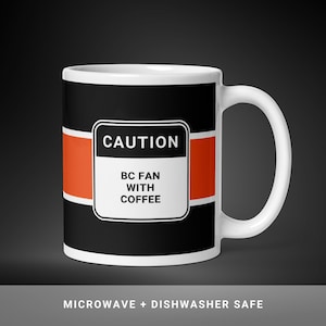 Può includere: Tazza in ceramica bianca con sfondo nero e striscia arancione. La tazza presenta il testo "CAUTION BC FAN WITH COFFEE" in un rettangolo bianco. La tazza è adatta al microonde e alla lavastoviglie.