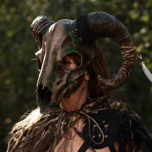 May include: A ram skull mask featuring large curved horns, textured brown bone-like surfaces, and patches of green moss. The mask is adorned with a white feather tied to one horn with twine, paired with a distressed brown burlap cape and black straps.