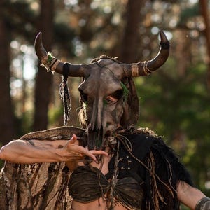 Könnte beinhalten: Eine gehörnte Tierschädelmaske in dunklen Brauntönen mit texturierten Oberflächen und umwickelten Garnakzenten an den Hörnern. Die Maske wird mit einem dunklen, abgenutzten Kleidungsstück kombiniert, das mit grobem Gewebe und dunklen Fransen versehen ist.