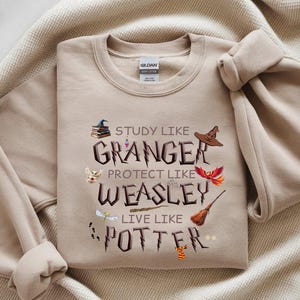 May include: Beige sweatshirt with the text "Study like Granger, Protect like Weasley, Live like Potter" and themed graphics. The design includes a stack of books, a sorting hat, a phoenix, a broom, and other elements.