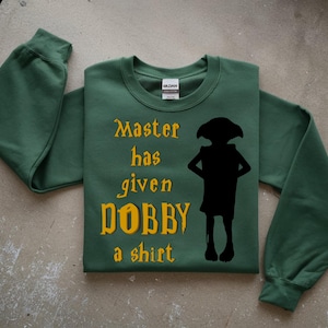May include: Green sweatshirt with the text "Master has given Dobby a shirt" in gold lettering. A black silhouette of Dobby the house-elf is also featured. The sweatshirt is folded on a light gray surface.