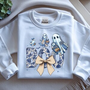 Op de afbeelding: Witte sweatshirt met een Halloween-thema ontwerp. Het woord "BOO" is geschreven in een blauw bloemenpatroon, met een beige strik. Drie spookfiguren met blauwe bloemenpatronen en pompoen-tassen staan boven het woord.