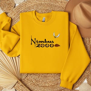 Könnte beinhalten: Senfgelbes Sweatshirt mit dem Text "Nimbus 2000" in schwarzer Schrift, einem goldenen Schnatz und einer braunen Besen-Grafik. Das Sweatshirt hat lange Ärmel und einen Rundhalsausschnitt.