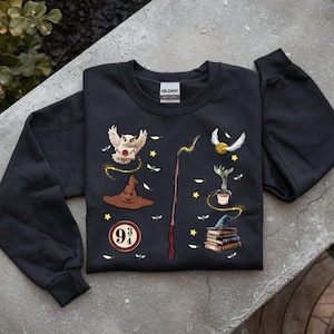 Puede incluir: Sudadera negra de manga larga con varios gráficos temáticos de Harry Potter. El diseño incluye un búho, el sombrero seleccionador, una varita, la snitch dorada, una planta, una pila de libros y el logo de la plataforma 9 3/4. Los gráficos son en tonos marrón, dorado y blanco.