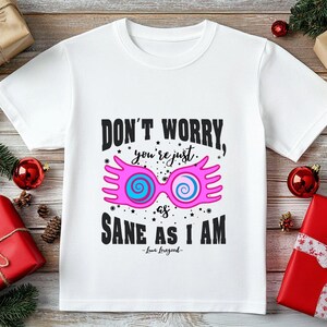 May include: White t-shirt with the text "DON'T WORRY, you're just as SANE AS I AM" and a graphic of pink and blue glasses. The shirt is surrounded by Christmas decorations, including wrapped gifts and ornaments.