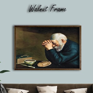 Op de afbeelding: Een ingelijst kunstwerk dat een oude man met een witte baard in gebed afbeeldt. Het schilderij, in een walnoten lijst, toont een tafel met een kom, brood en een boek. De tekst "Walnut Frame" staat boven het kunstwerk.