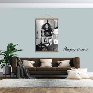 May include: A black and white hanging canvas print featuring a person with long hair sitting on a toilet, holding a cigarette and a glass. The artwork is mounted on a wooden frame with a string for hanging, displayed on a wall.