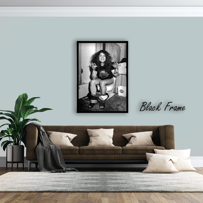 May include: A black and white framed photograph depicting a person with long hair sitting on a toilet, holding a glass and a cigarette. The person wears a dark t-shirt and shorts. The image is enclosed in a simple black frame, displayed on a wall.