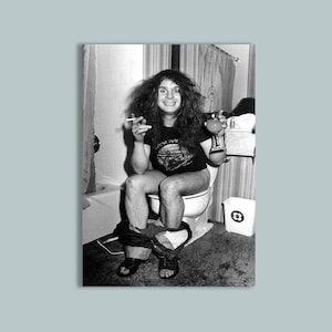 May include: A black and white photograph featuring a person with long dark hair wearing a black t-shirt with a graphic print. The person is seated on a toilet, holding a cigarette in one hand and a glass of liquid in the other, wearing dark sandals.