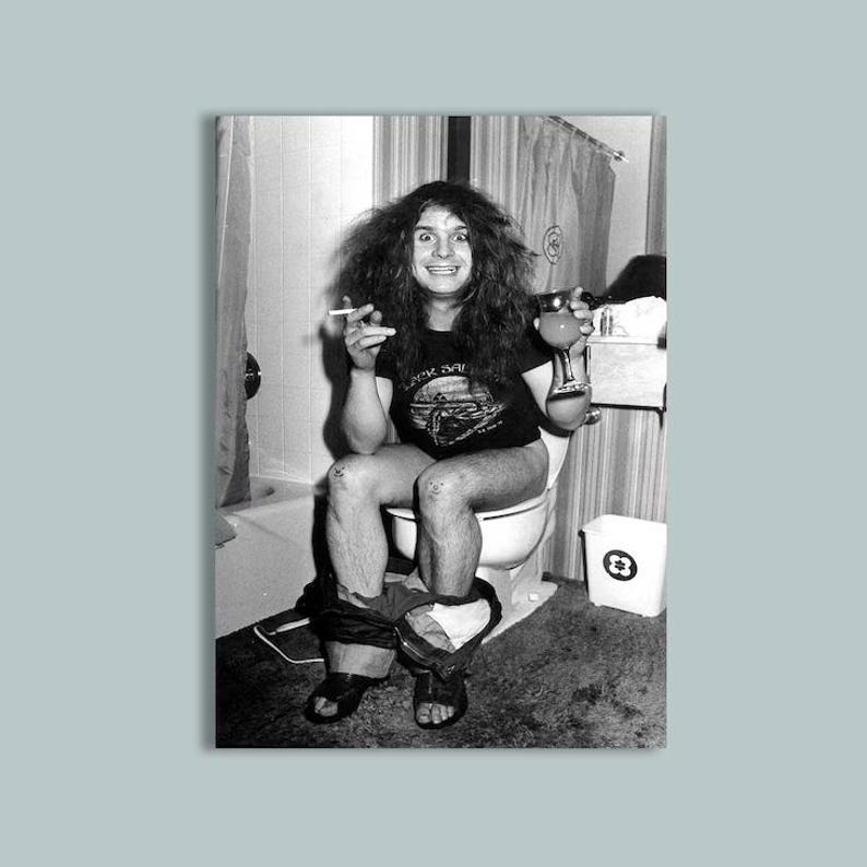 May include: A black and white photograph featuring a person with long dark hair wearing a black t-shirt with a graphic print. The person is seated on a toilet, holding a cigarette in one hand and a glass of liquid in the other, wearing dark sandals.