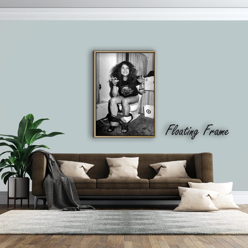 May include: A black and white framed wall art print featuring a person with long hair sitting on a toilet, holding a glass and a cigarette, wearing a t-shirt and shorts. The artwork is presented in a wooden floating frame, capturing a candid moment in a bathroom setting with a waste bin visible nearby.