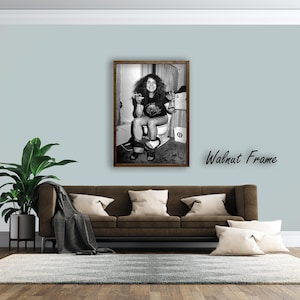 May include: A black and white framed photograph featuring a person with long hair sitting on a toilet, wearing a t-shirt and sandals, holding a cigarette and a glass. The image is enclosed in a dark brown wooden walnut frame.