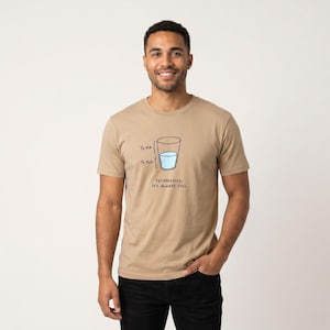 May include: A tan short-sleeve t-shirt featuring a graphic of a glass half-filled with blue water. The graphic includes labels for 1/2 air and 1/2 H2O, with the text below the glass reading, "TECHNICALLY, IT'S ALWAYS FULL" in black lettering.