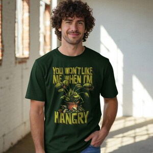 May include: A dark green short-sleeve t-shirt featuring a graphic of a Venus flytrap in a terracotta pot surrounded by skulls. The text above the plant reads "YOU WON'T LIKE ME WHEN I'M" and the text below the plant reads "HANGRY" in yellow.