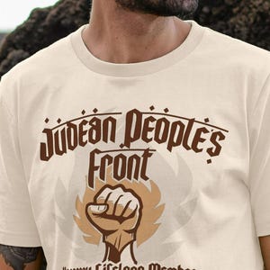 May include: A cream-coloured t-shirt with the brown text "Judean People's Front" and a clenched fist graphic. The shirt also features the text "Honorary Lifelong Member".