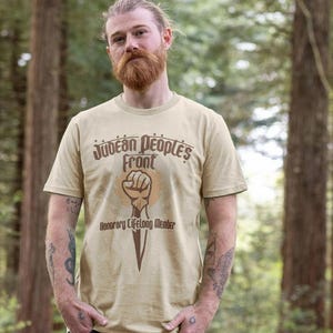 May include: A beige short-sleeve t-shirt featuring a brown graphic of a raised fist holding a dagger. The text reads Judean People's Front and Honorary Lifelong Member in a stylised font. The shirt has a crew neckline and a standard fit.