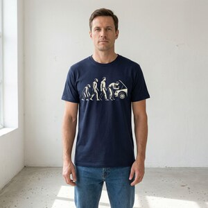 May include: Navy blue t-shirt with a white graphic illustrating human evolution, ending with a person inspecting a car engine. The design shows the progression from ape to mechanic. The shirt is worn by a person.