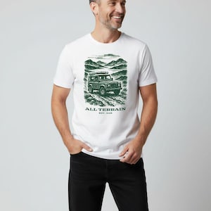 May include: White t-shirt featuring a green illustration of a 4x4 vehicle on a dirt track, with a mountain scene. The text "ALL TERRAIN EST. 1948" is printed below. The person is wearing black jeans.