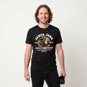 May include: Black t-shirt with a Jawa Java graphic. The design features a cartoon Jawa character holding a coffee cup, with text that reads "Extra Caffeinated: The Strongest in the Galaxy." The shirt is worn by a person holding a black mug.